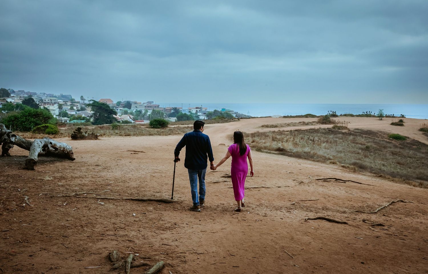 Couple holding hands walking on sandy coastal path under cloudy sky, woman in pink dress, man with walking stick.