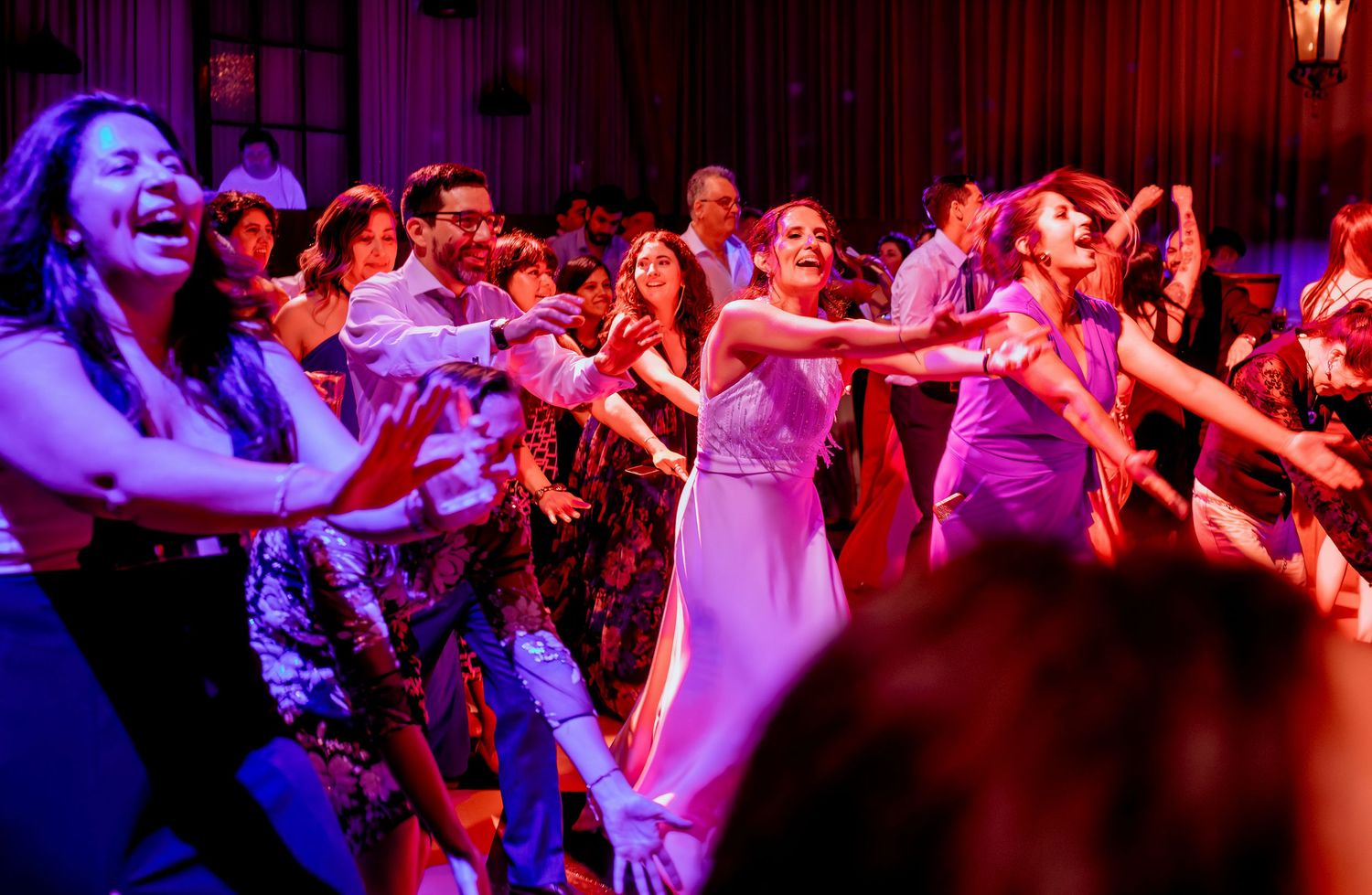 Joyful wedding guests in colorful attire laugh and dance together on the reception dance floor.