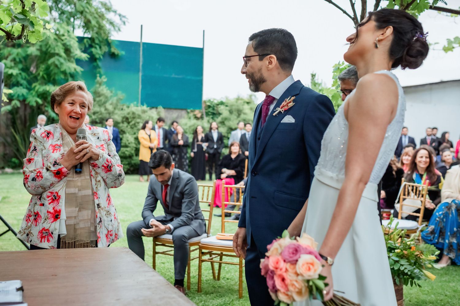 Officiant laughs with bride and groom during joyful outdoor wedding ceremony.