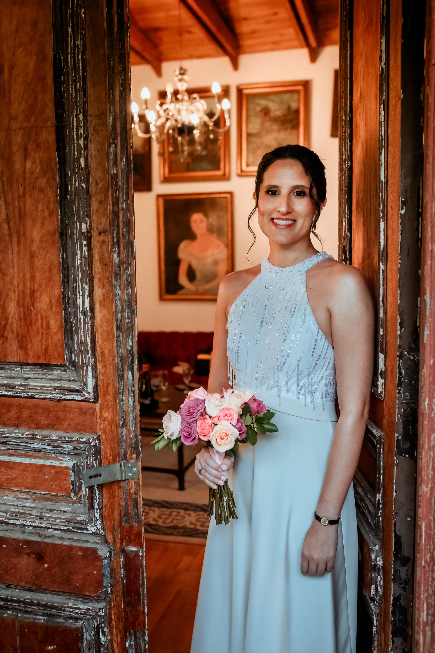 Elegant bride in light blue halter gown holding pink rose bouquet standing in rustic wooden doorway.