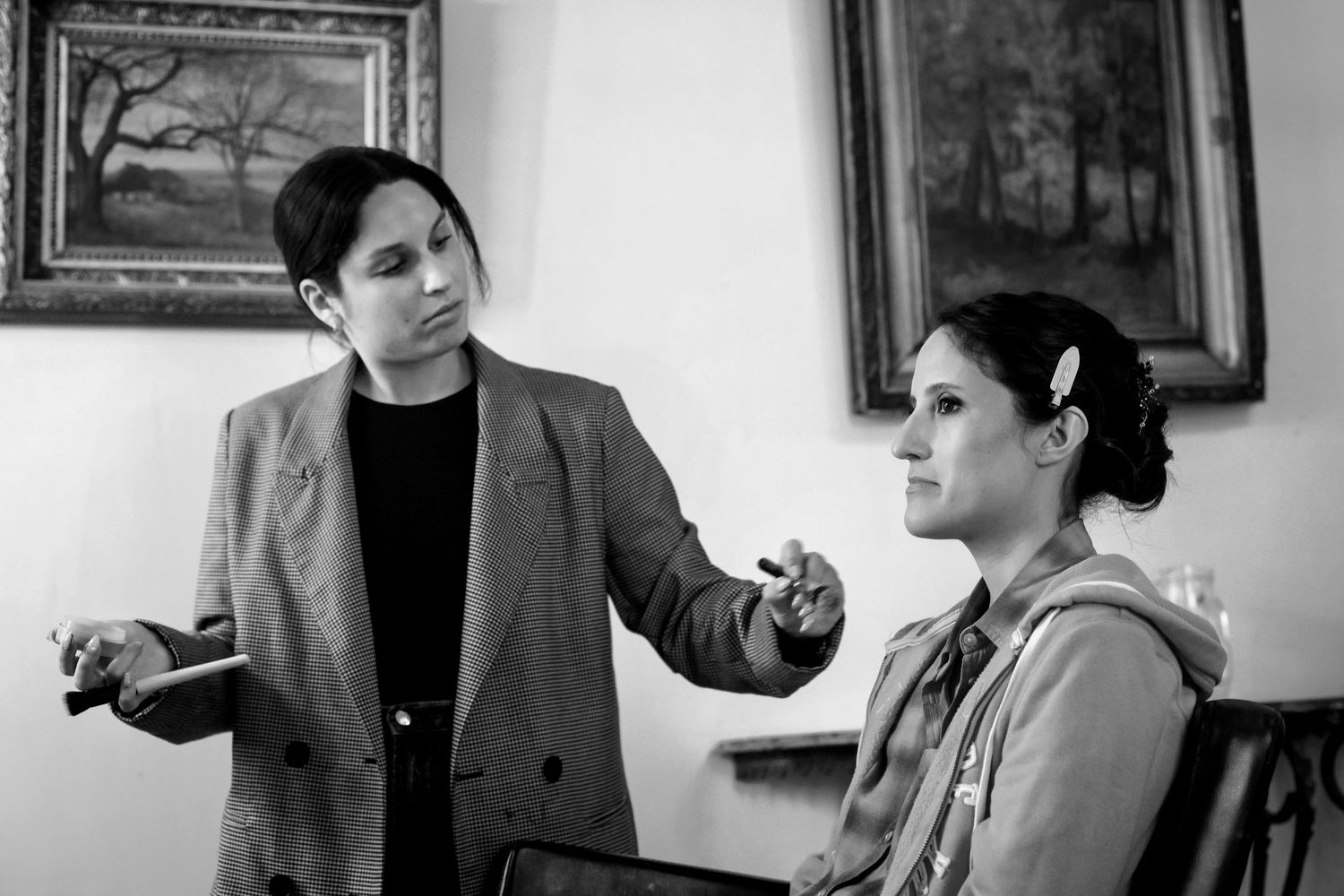 Black and white photo of makeup artist applying cosmetics to seated woman in studio.
