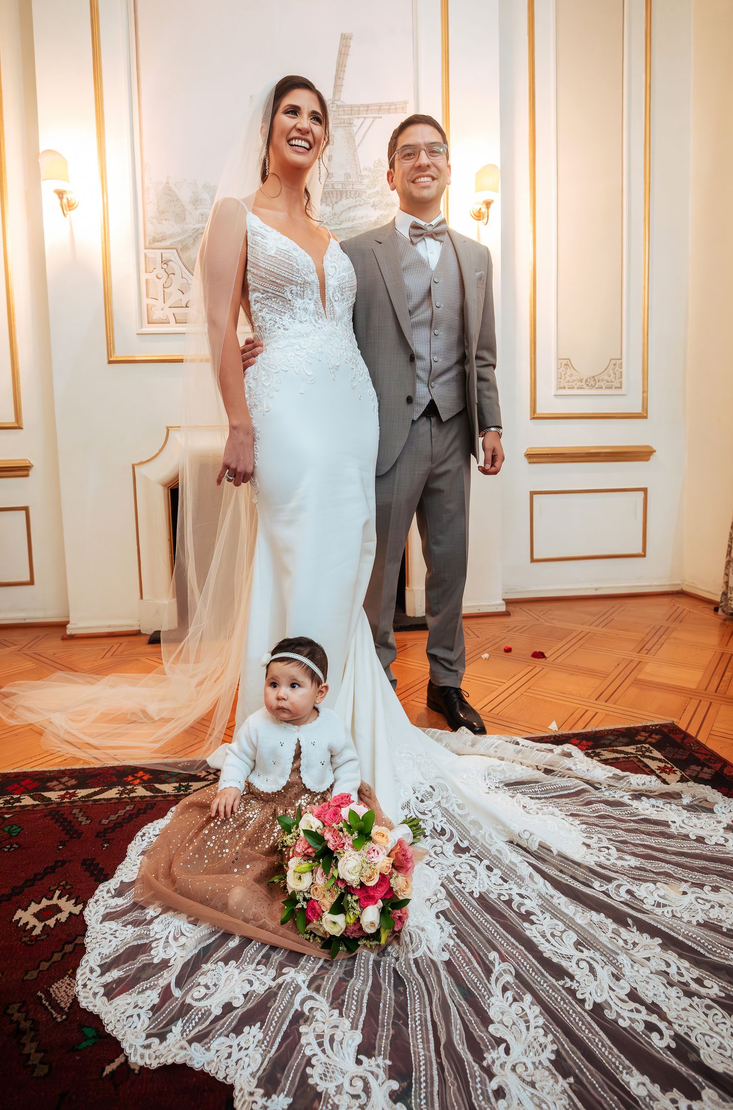 Bride in white mermaid gown and groom in gray suit pose with a baby sitting on the dress train holding a bouquet.
