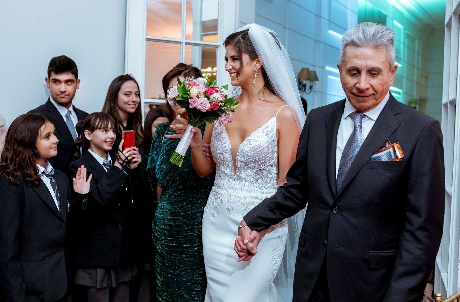 Bride in white lace gown holding bouquet walks with older man as family watches at elegant wedding venue.