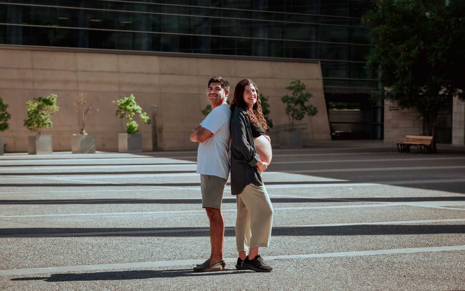 Pregnant couple standing back-to-back on a striped pavement plaza beside a contemporary stone building.