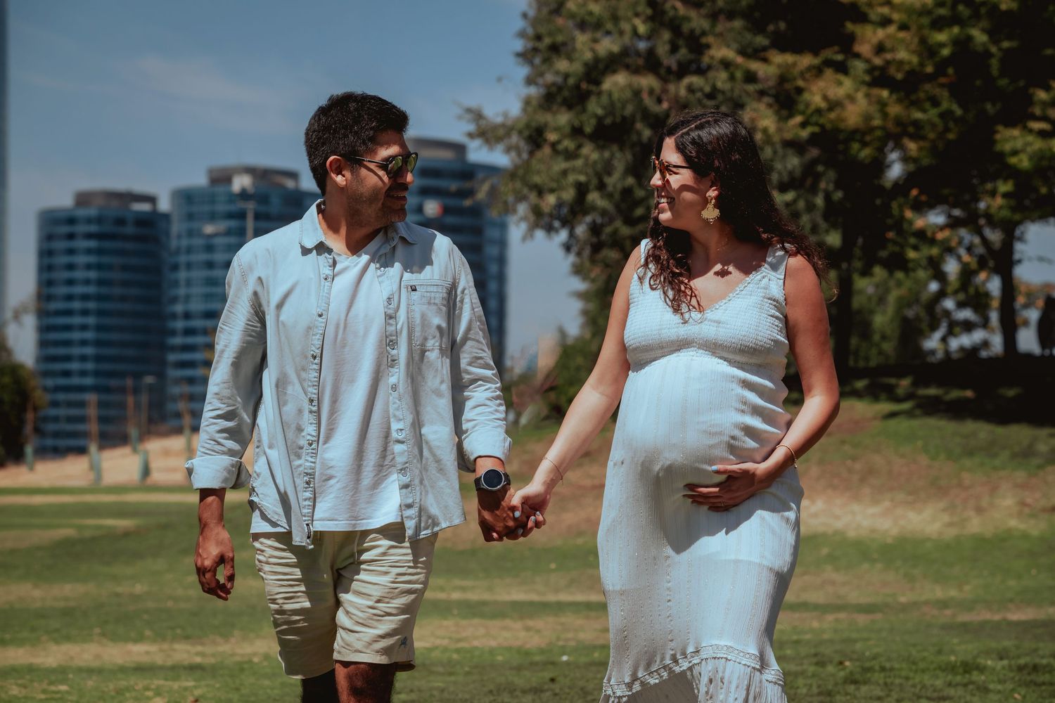 Pregnant couple walking hand in hand through a sunny park with city buildings in the background.