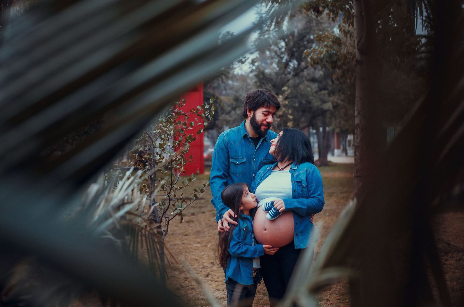 Couple sharing a tender moment outdoors, pregnant woman and bearded man framed by leaves.
