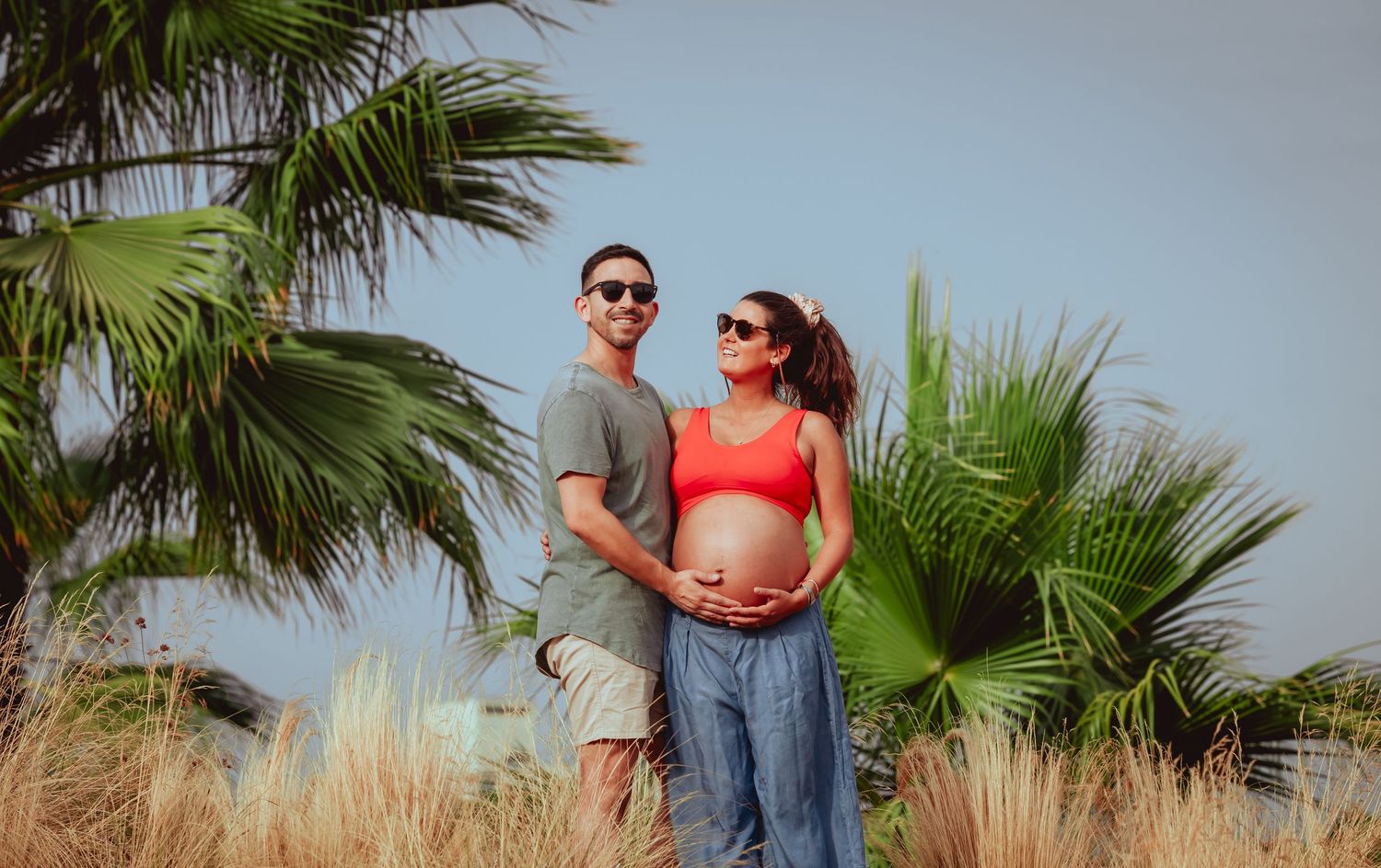 Couple posing outdoors with palm trees, pregnant woman in red top holding baby bump with partner beside her.