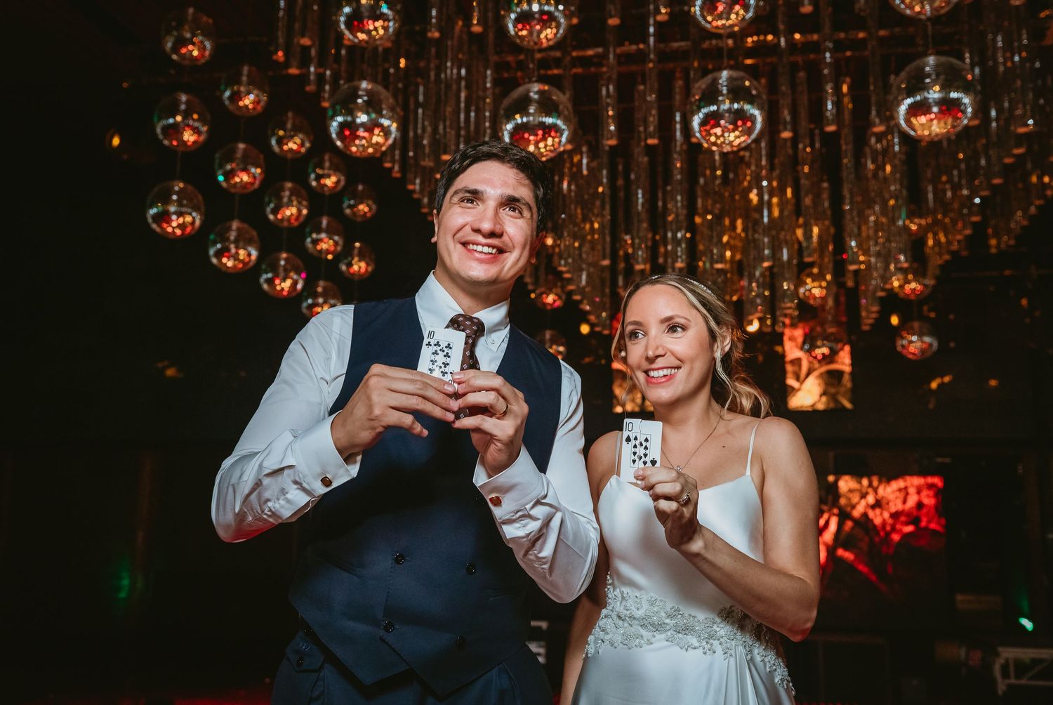 Bride and groom smile holding playing cards at their wedding reception with glowing sphere decorations above.