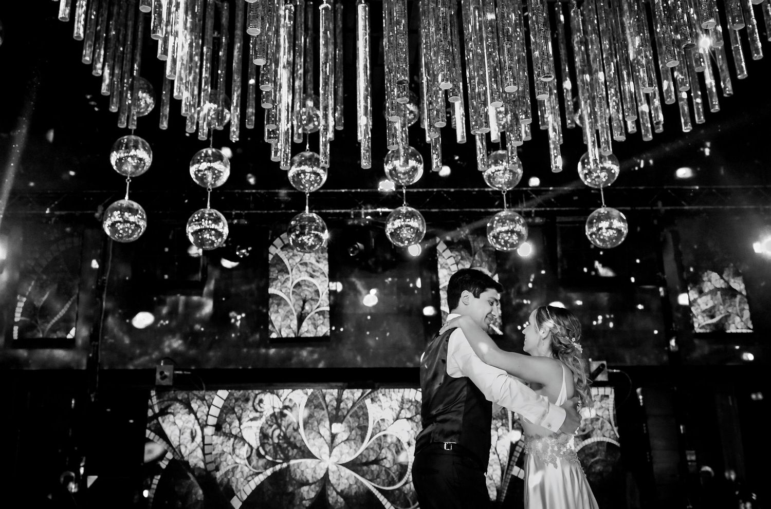 Black and white photo of bride and groom sharing first dance under crystal chandelier at elegant wedding venue.