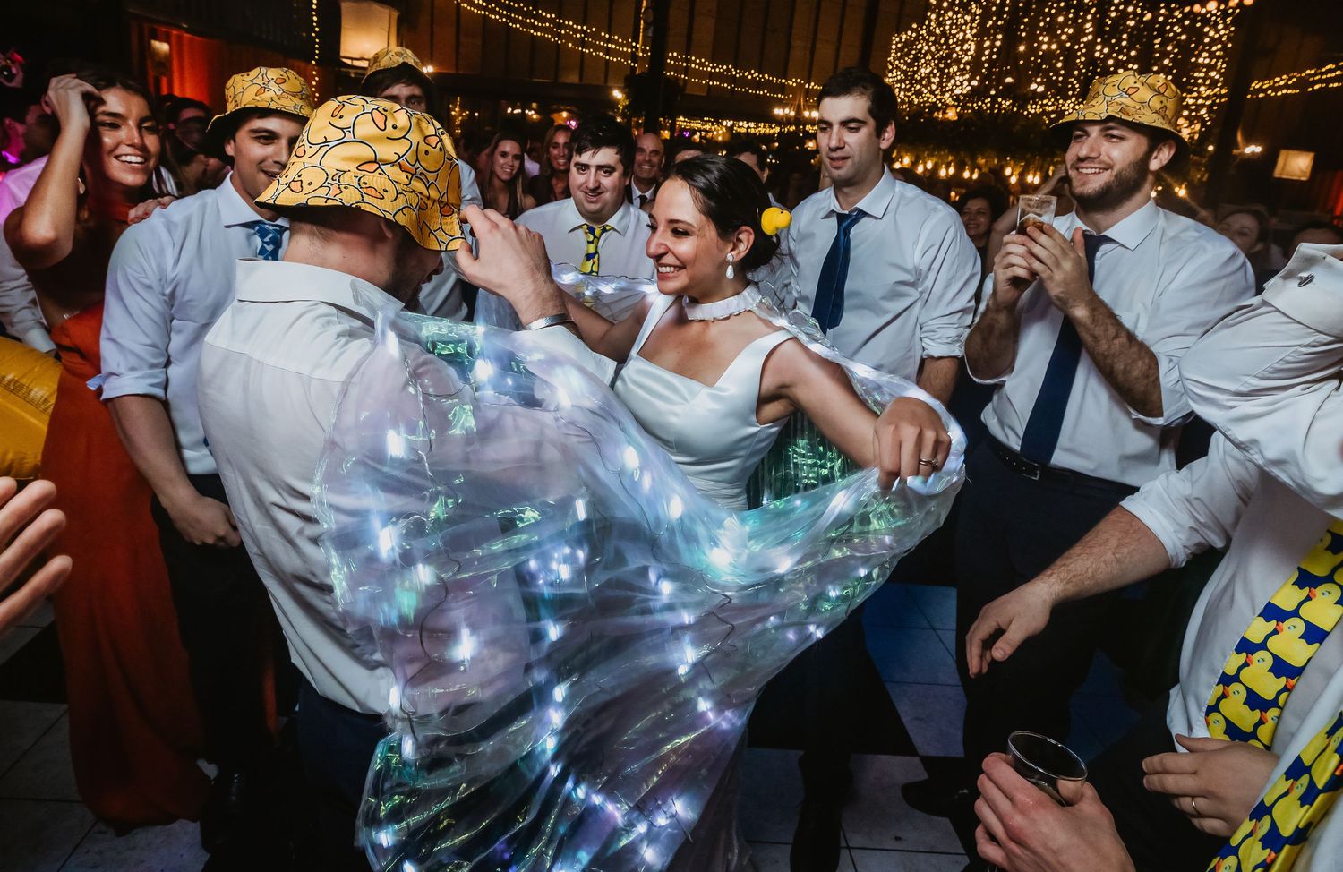 Bride and groom dancing joyfully at wedding reception with guests celebrating around them, wearing festive gold hats.