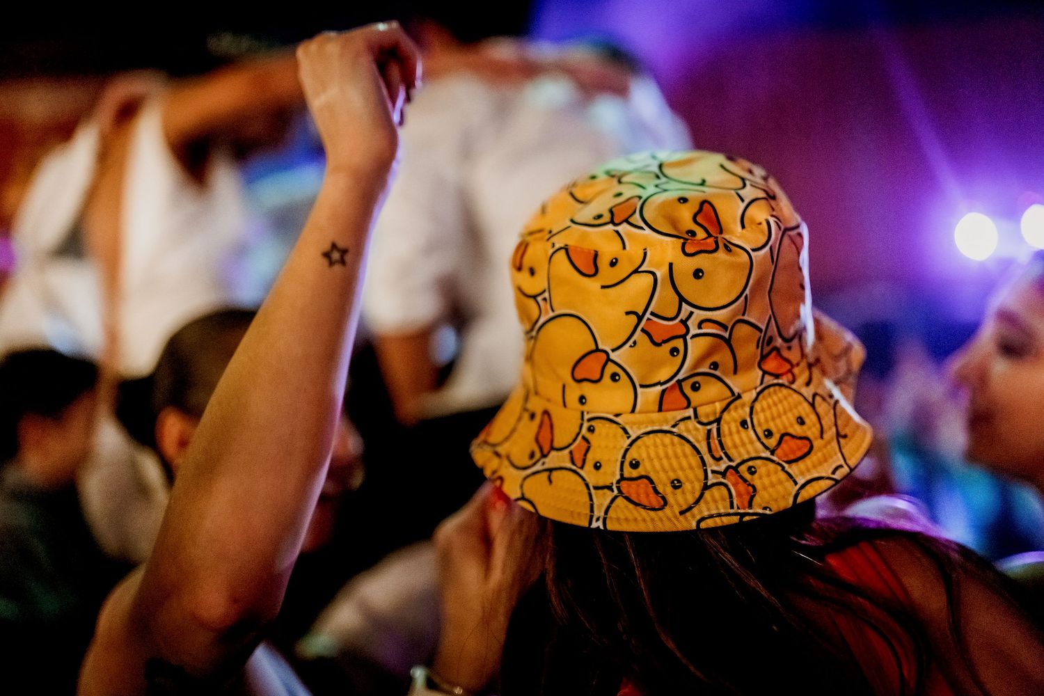 Person wearing a yellow bucket hat with duck pattern at a colorful music festival concert event.