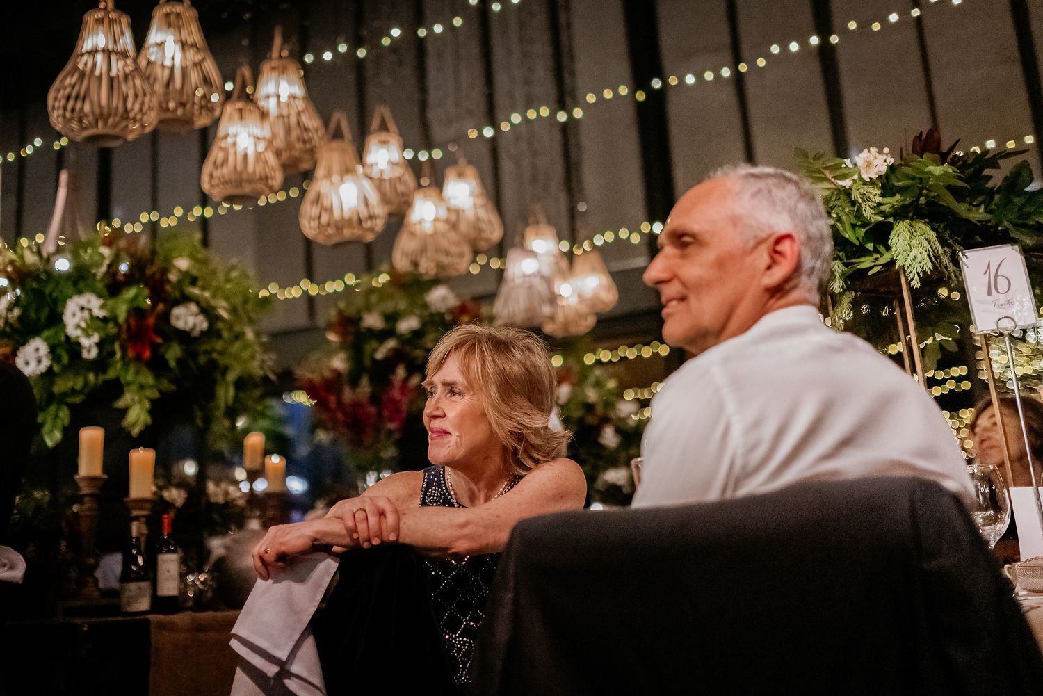 Elderly couple sitting together at an elegant wedding reception with warm string lights and floral decorations.