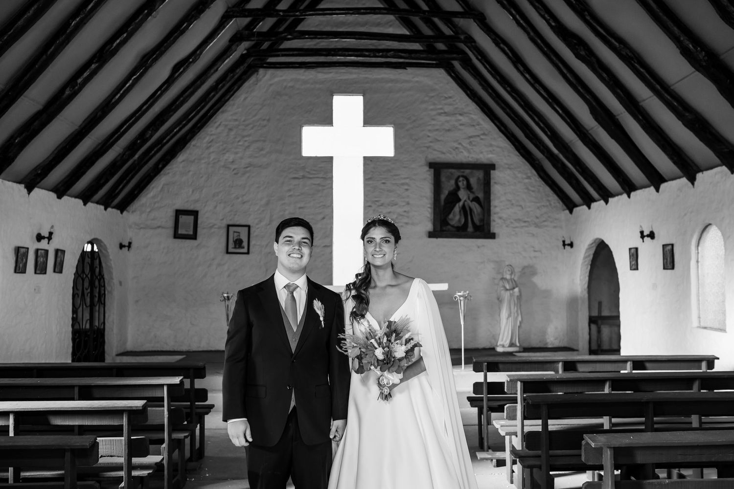 Black and white photo of a bride and groom standing together inside a small chapel with a glowing cross.
