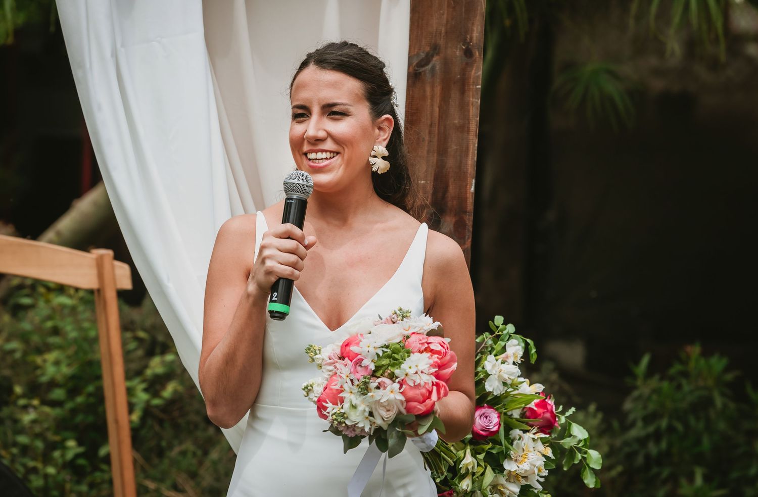 Smiling bride in white gown holds bouquet of pink roses and microphone at outdoor wedding ceremony.