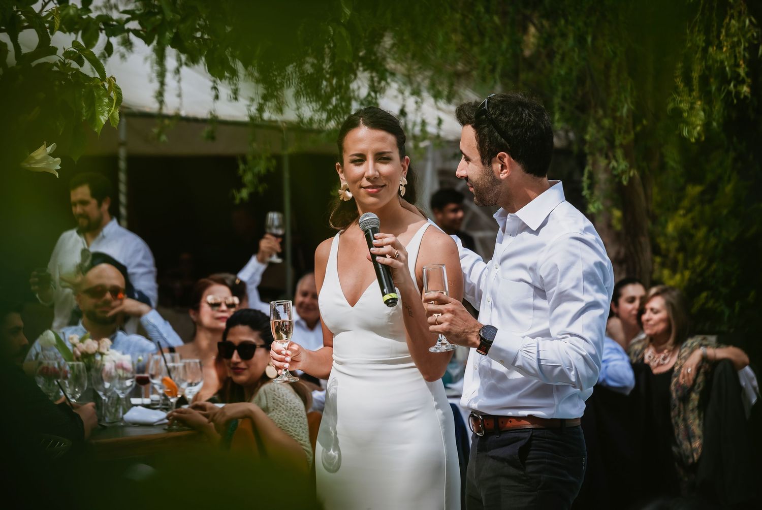 Bride and groom share a microphone during outdoor wedding reception toast, surrounded by guests among lush green trees.