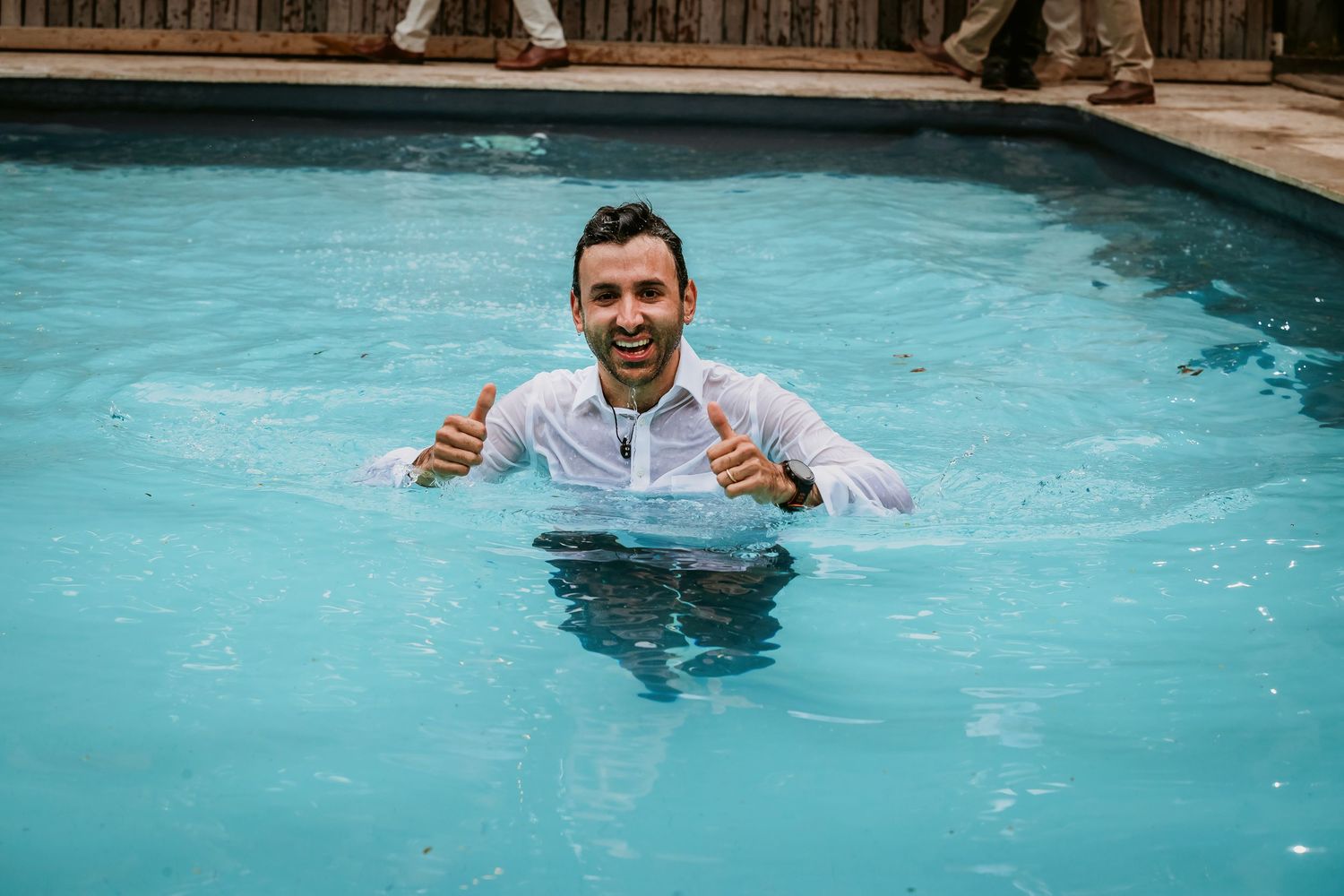 Man in white shirt giving thumbs up while standing in a swimming pool, smiling joyfully.