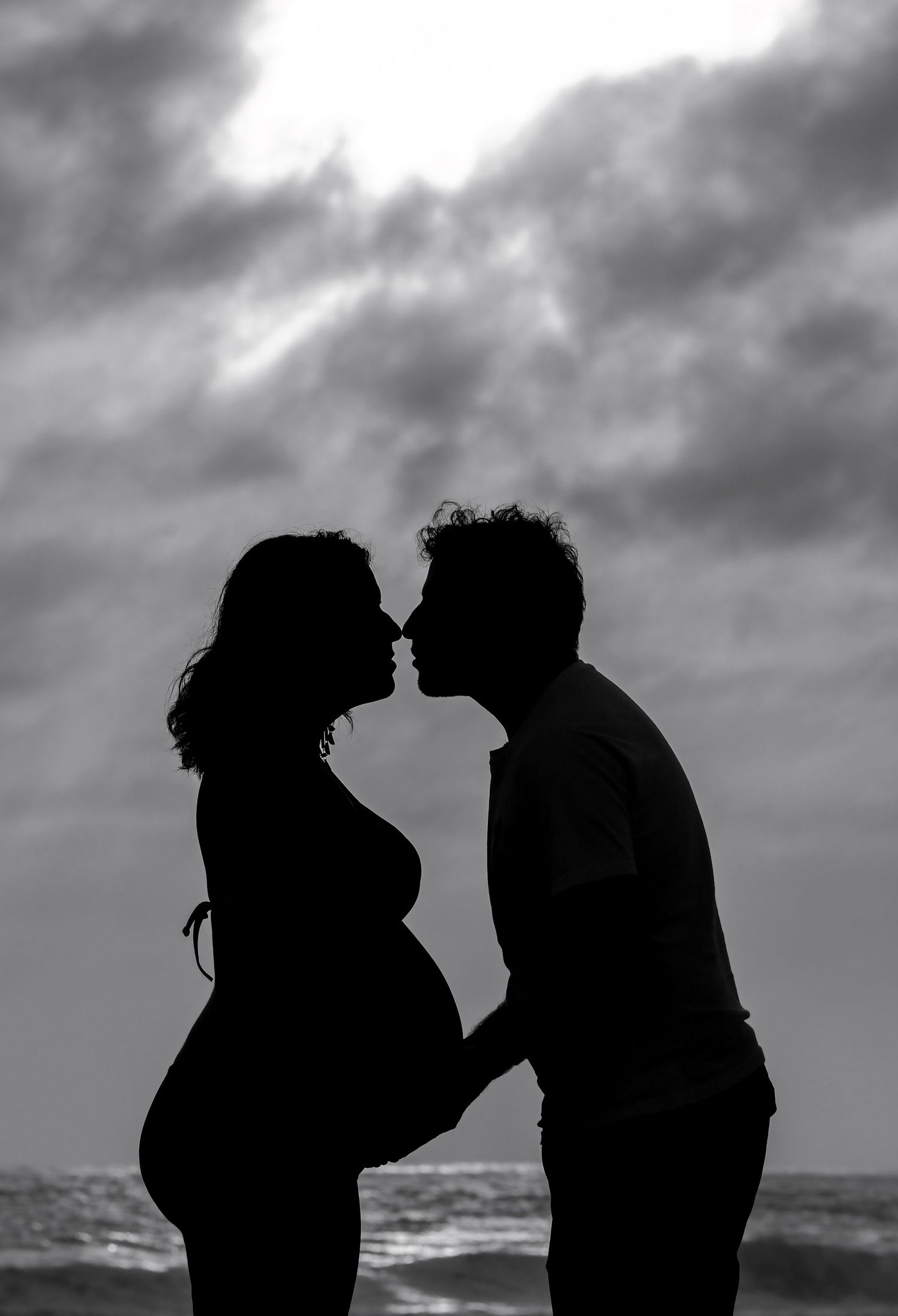Black and white silhouette of a pregnant couple kissing on a beach with dramatic cloudy sky backdrop.