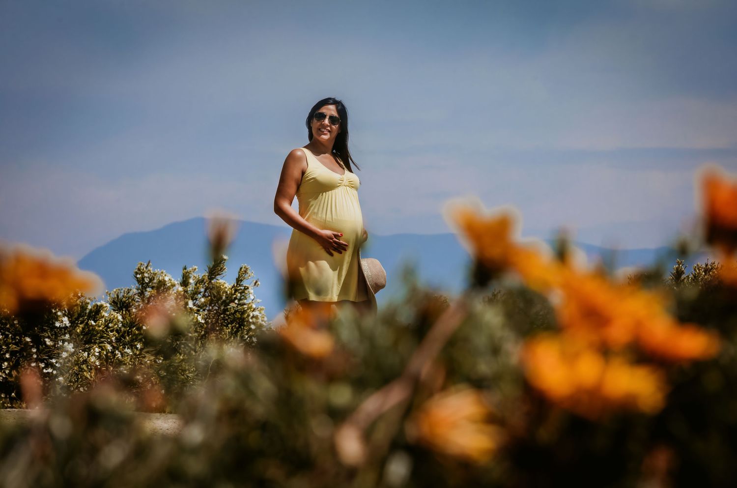 Pregnant woman in yellow dress standing among orange wildflowers with mountains in the background on a sunny day.