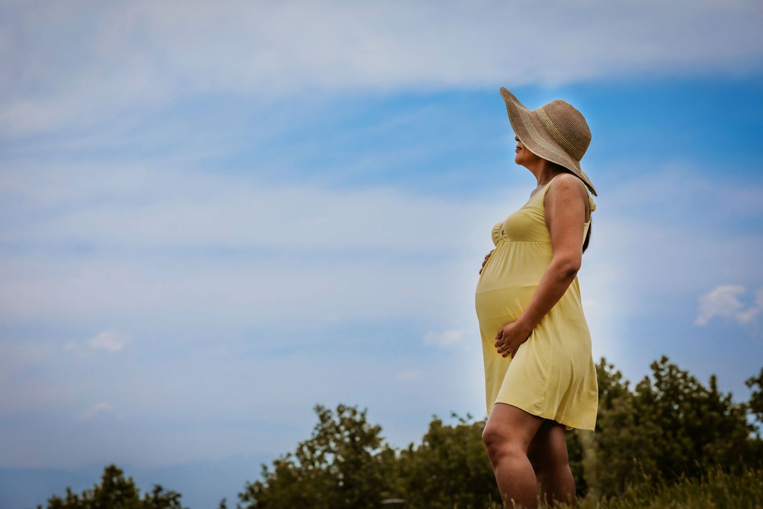 Pregnant woman in yellow dress and sun hat standing outdoors against a blue sky backdrop.