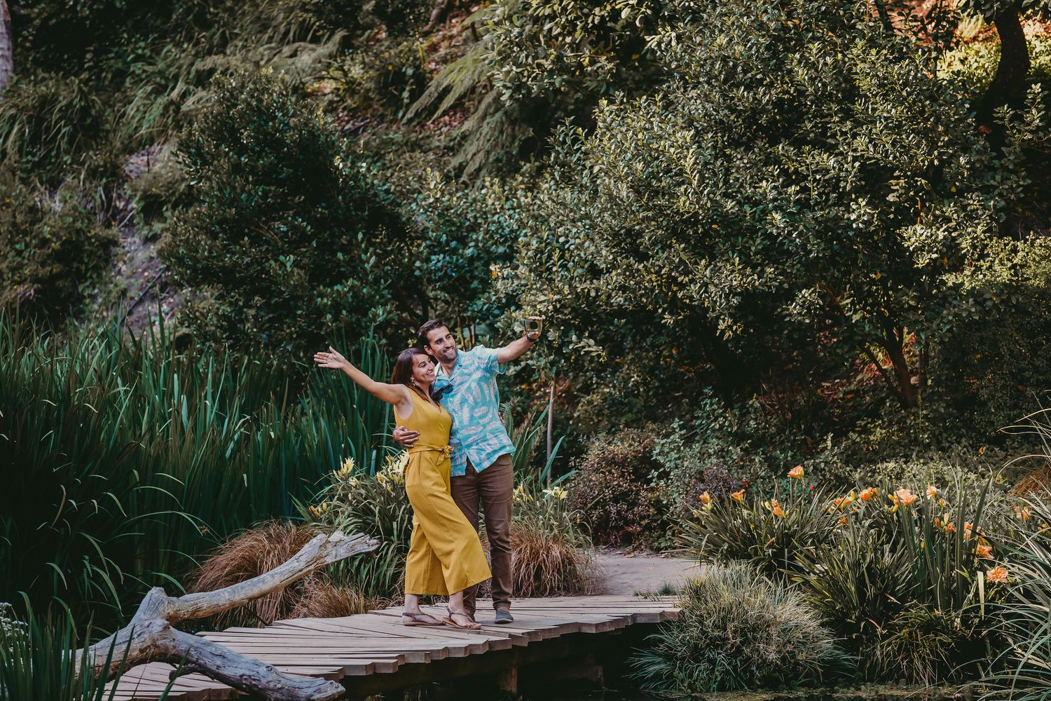 Joyful couple with arms outstretched standing on a wooden bridge surrounded by garden greenery.