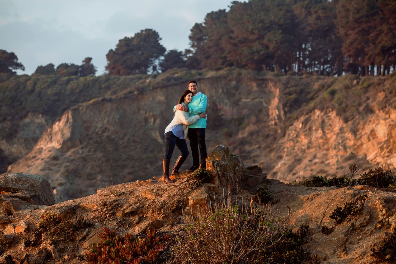 Romantic couple embracing on rugged coastal cliffs with warm golden light and pine trees in background.