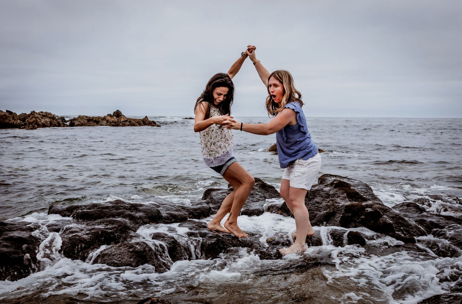 Two women laugh and dance together on wet coastal rocks with ocean waves splashing.