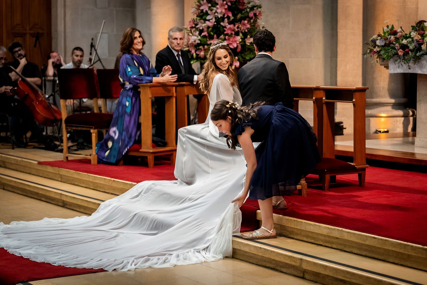 Bride in white gown with long veil has dress adjusted by flower girl at church altar during wedding ceremony.
