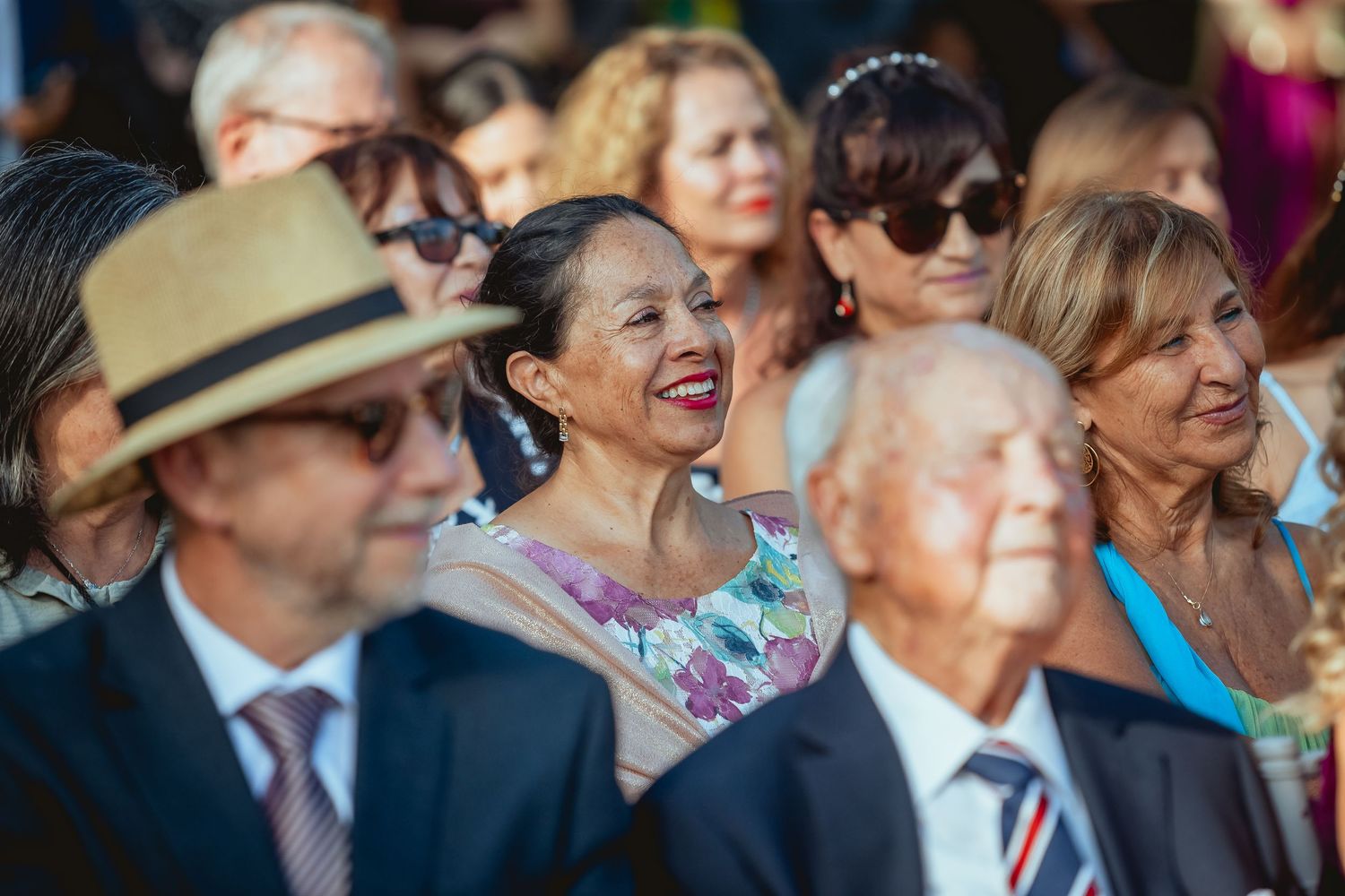 Joyful woman in floral dress laughs among smiling wedding guests seated at outdoor ceremony.