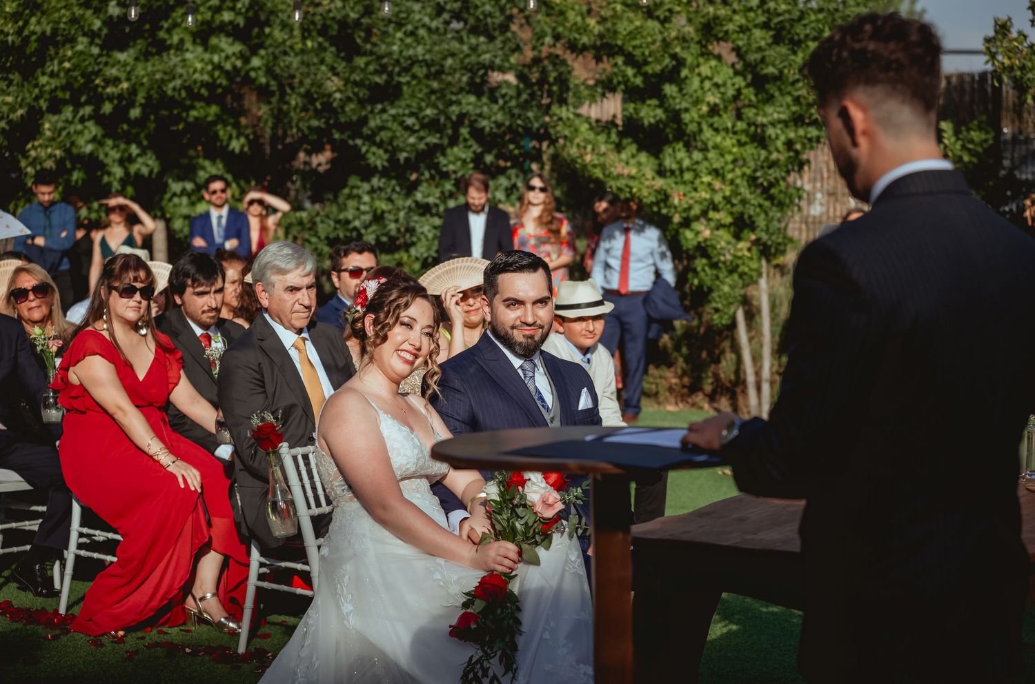 Bride smiles at groom during outdoor wedding ceremony while guests watch from white chairs.