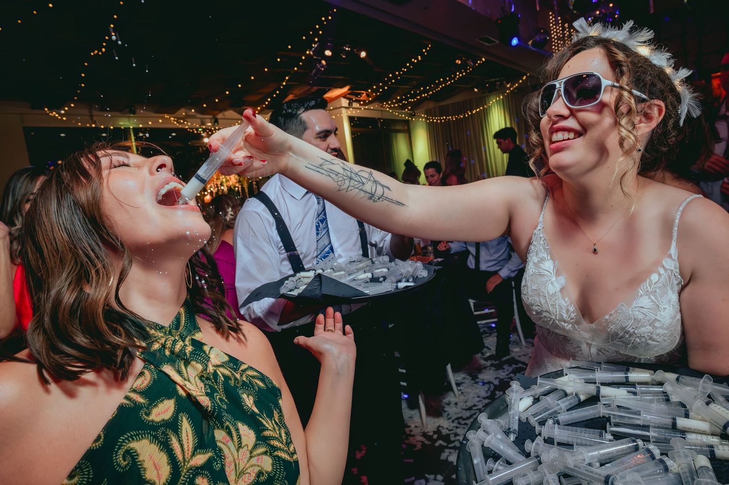 Bride in lace gown giving shot to guest in green dress at fun wedding reception.