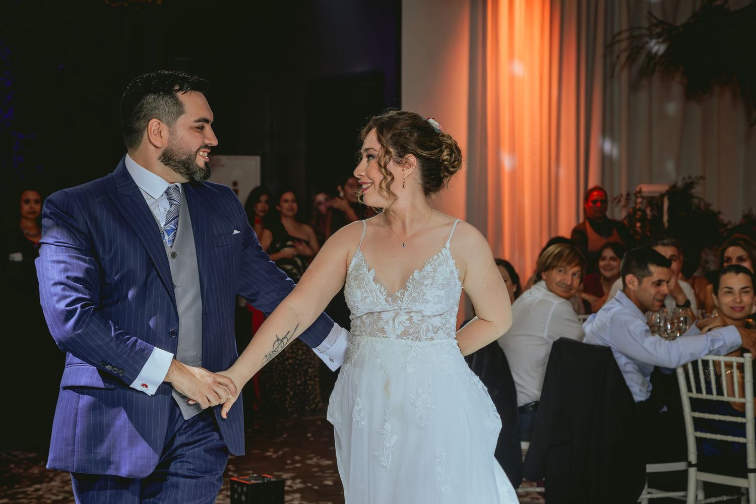 Smiling bride and groom share their first dance at elegant wedding reception with warm amber lighting.