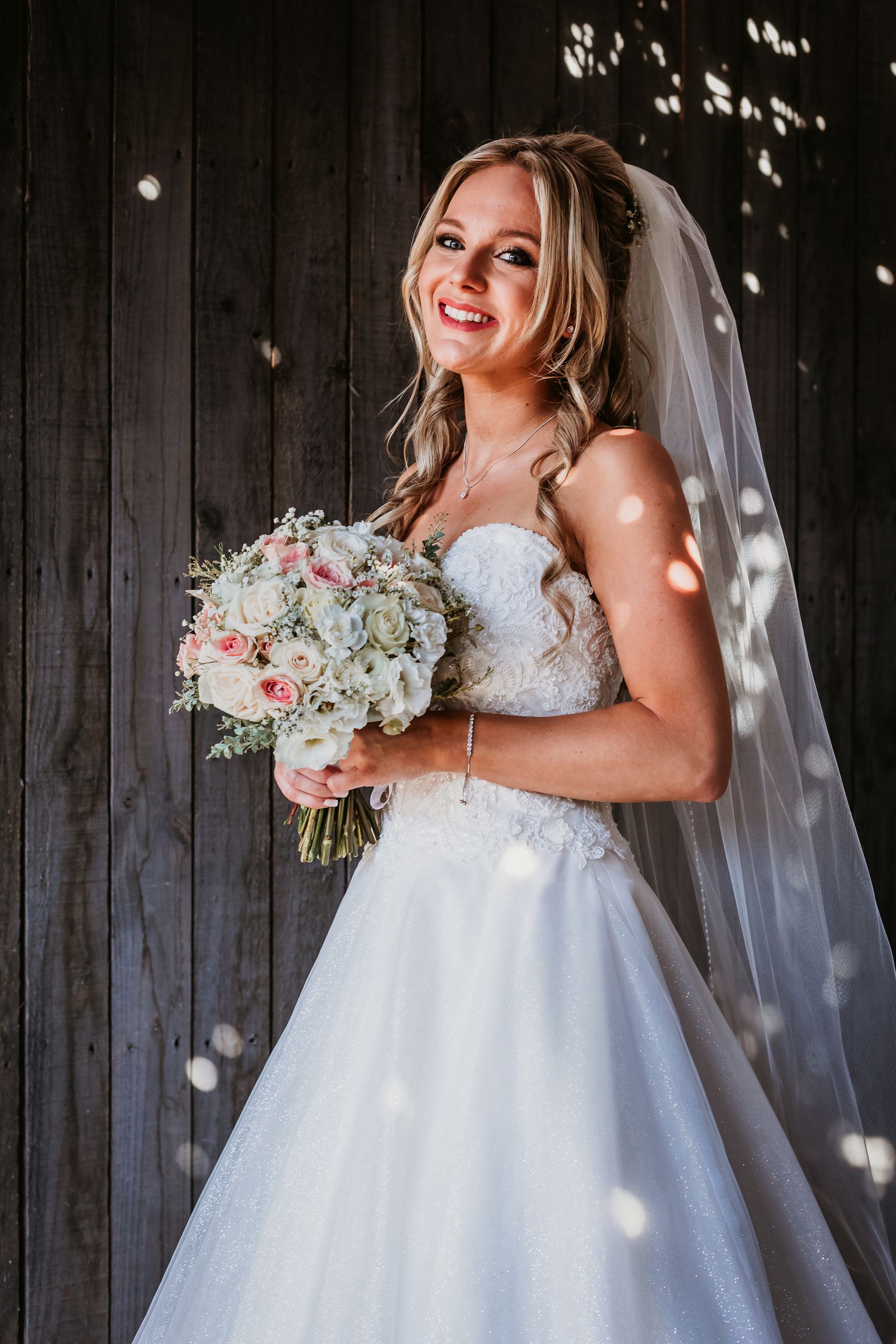 Smiling bride in a strapless lace wedding gown holds a bouquet against a dark wood wall.