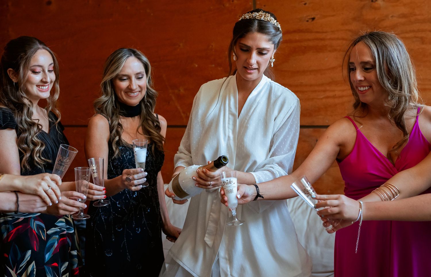 Bride in white robe pouring champagne for smiling bridesmaids in colorful dresses before the wedding ceremony.