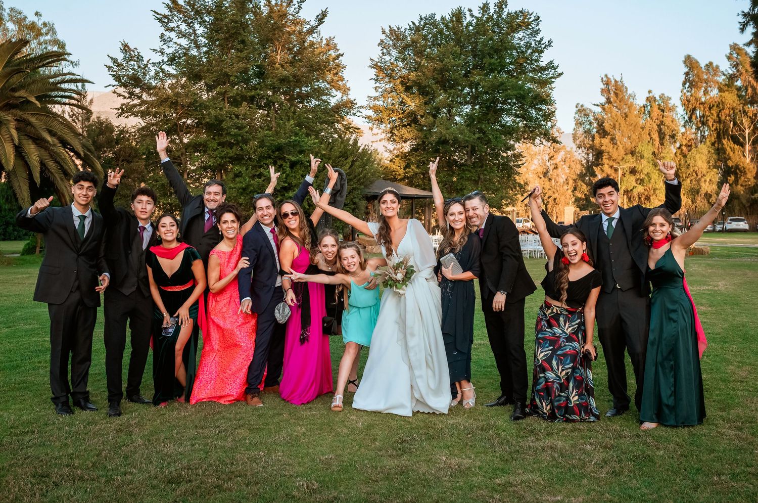 Large wedding party celebrates together with raised arms on a sunny green lawn with tall trees.