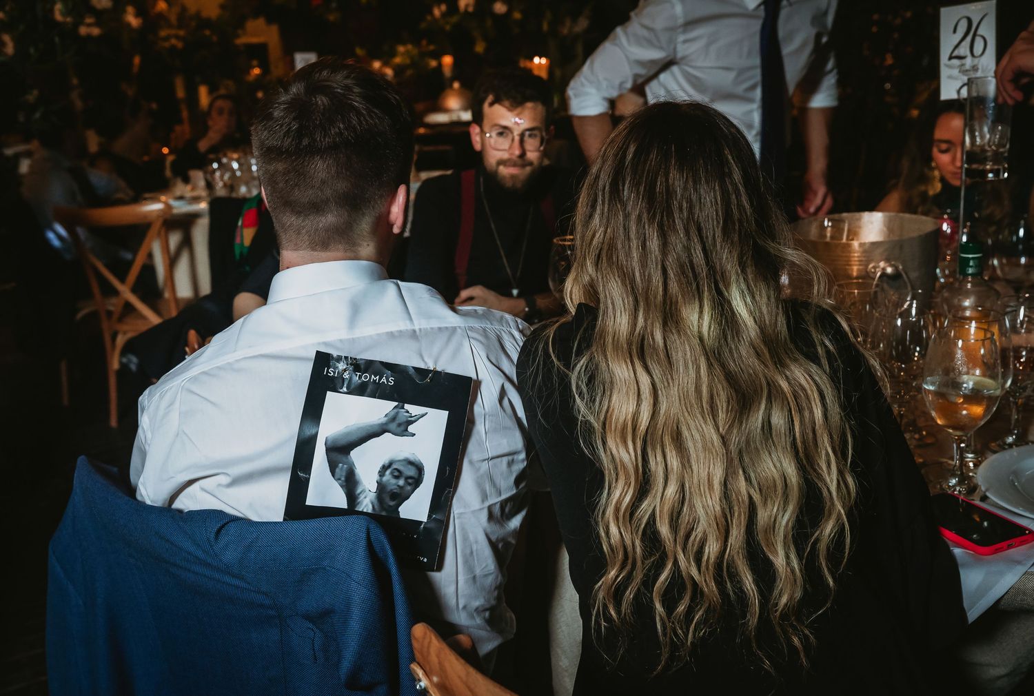 Guests at a formal dinner event, one wearing a graphic jacket, seated at a elegantly set table in dimly lit venue.