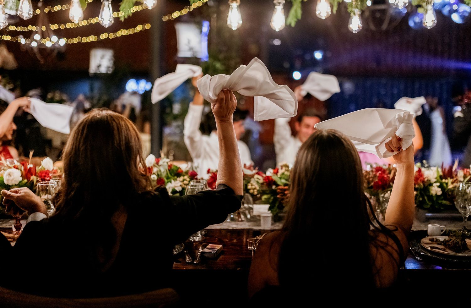 Guests wave white napkins at an elegant outdoor dinner party with floral centerpieces and string lights.