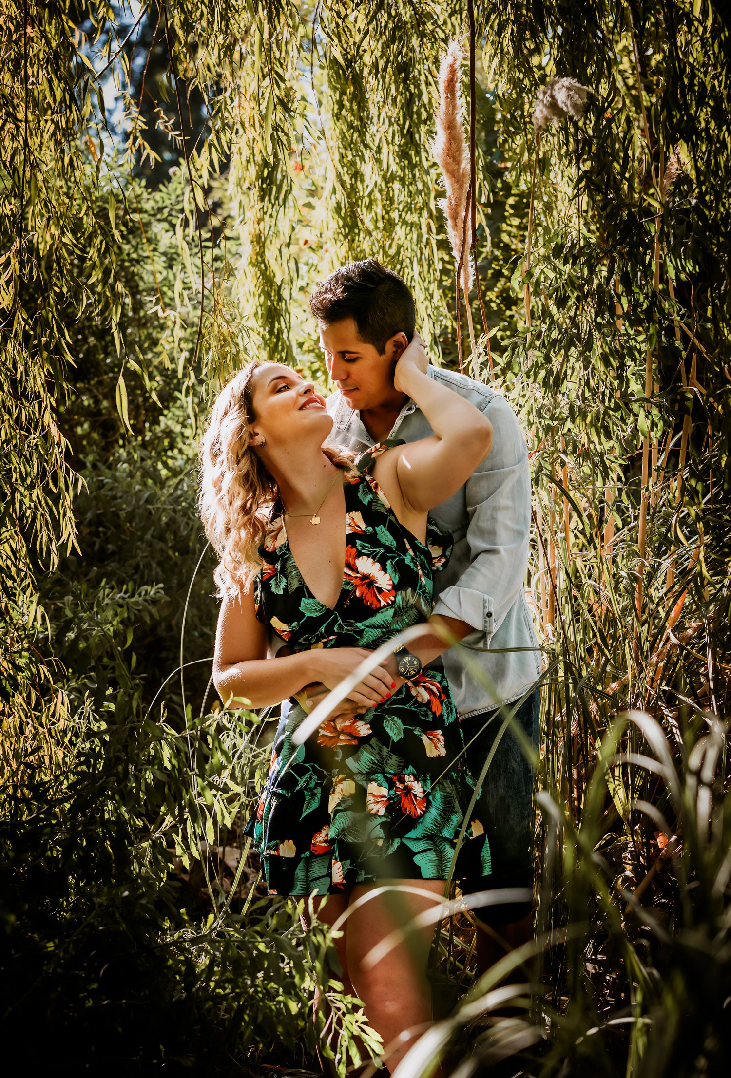 A couple embraces romantically under weeping willow branches, woman in floral dress, man in white shirt, surrounded by lush greenery.