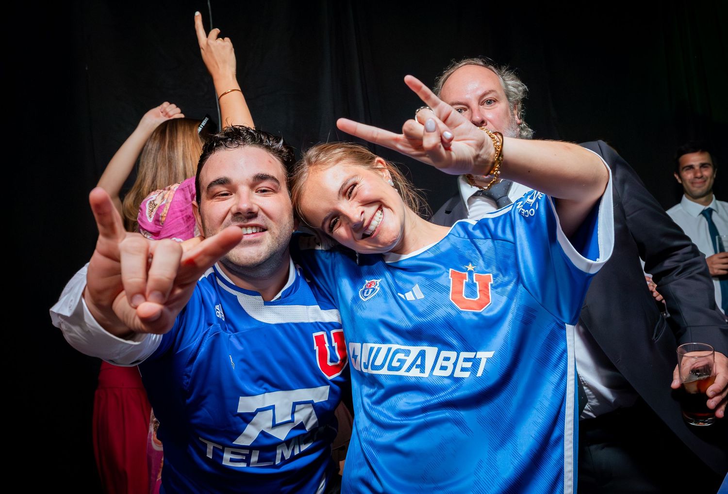 Two fans in blue Universidad de Chile jerseys pose making peace signs at a sports event.