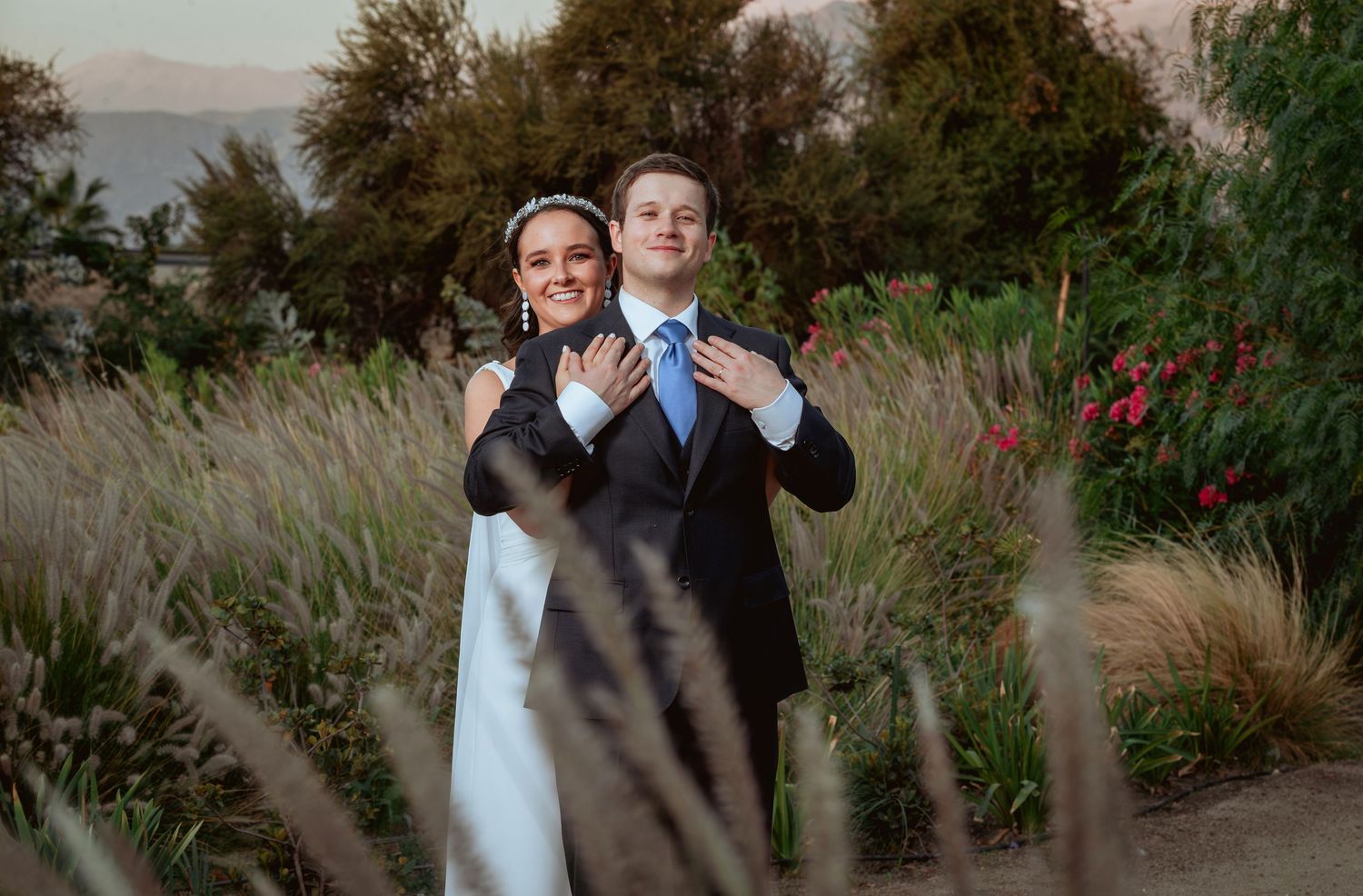 A bride and groom embrace in a lush garden setting at dusk, with mountains visible in the background.