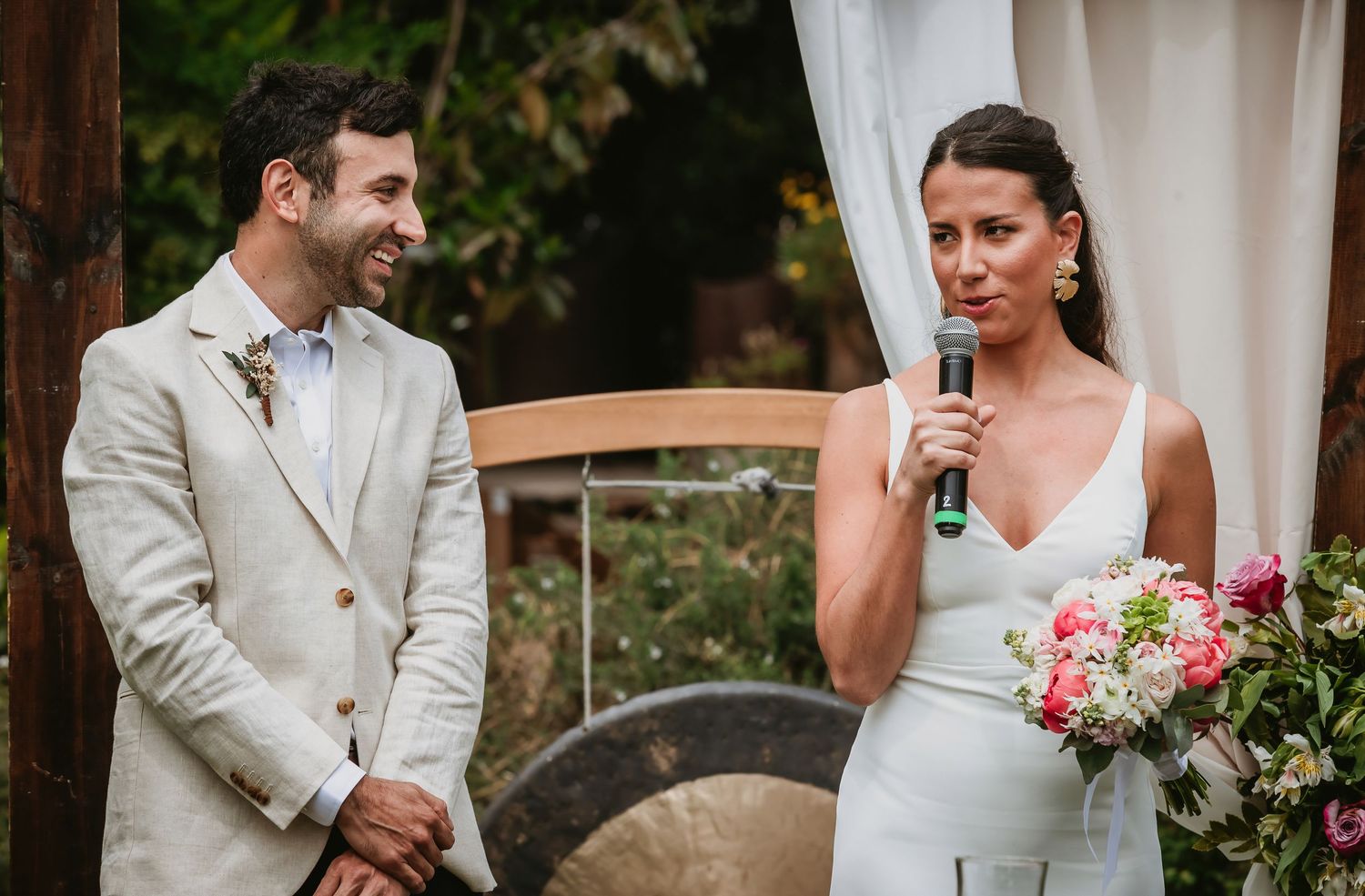 Bride speaks into microphone holding pink bouquet beside groom in beige suit at outdoor wedding ceremony.