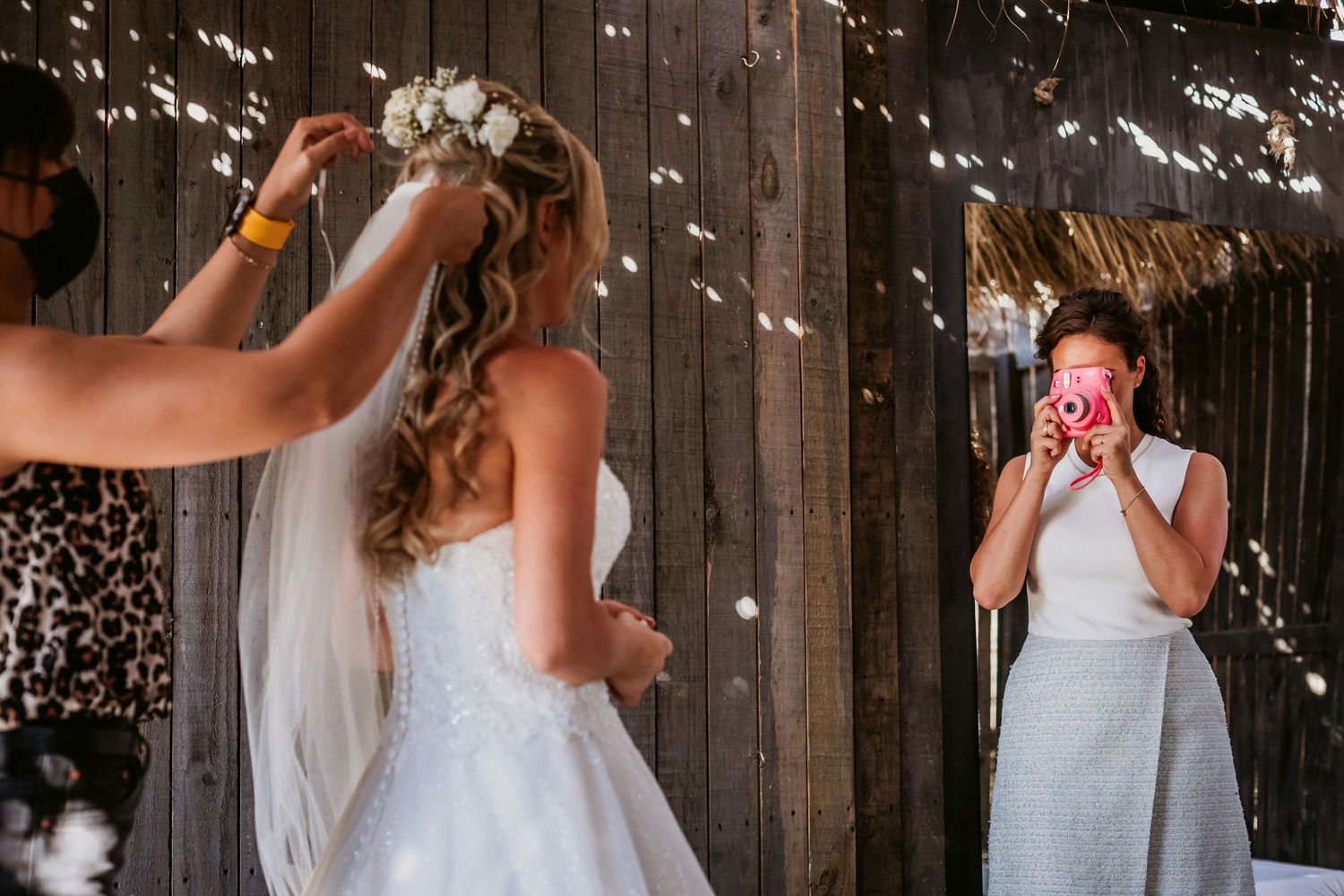 Wedding attendant adjusting bride's veil while guest photographs moment at rustic barn venue.