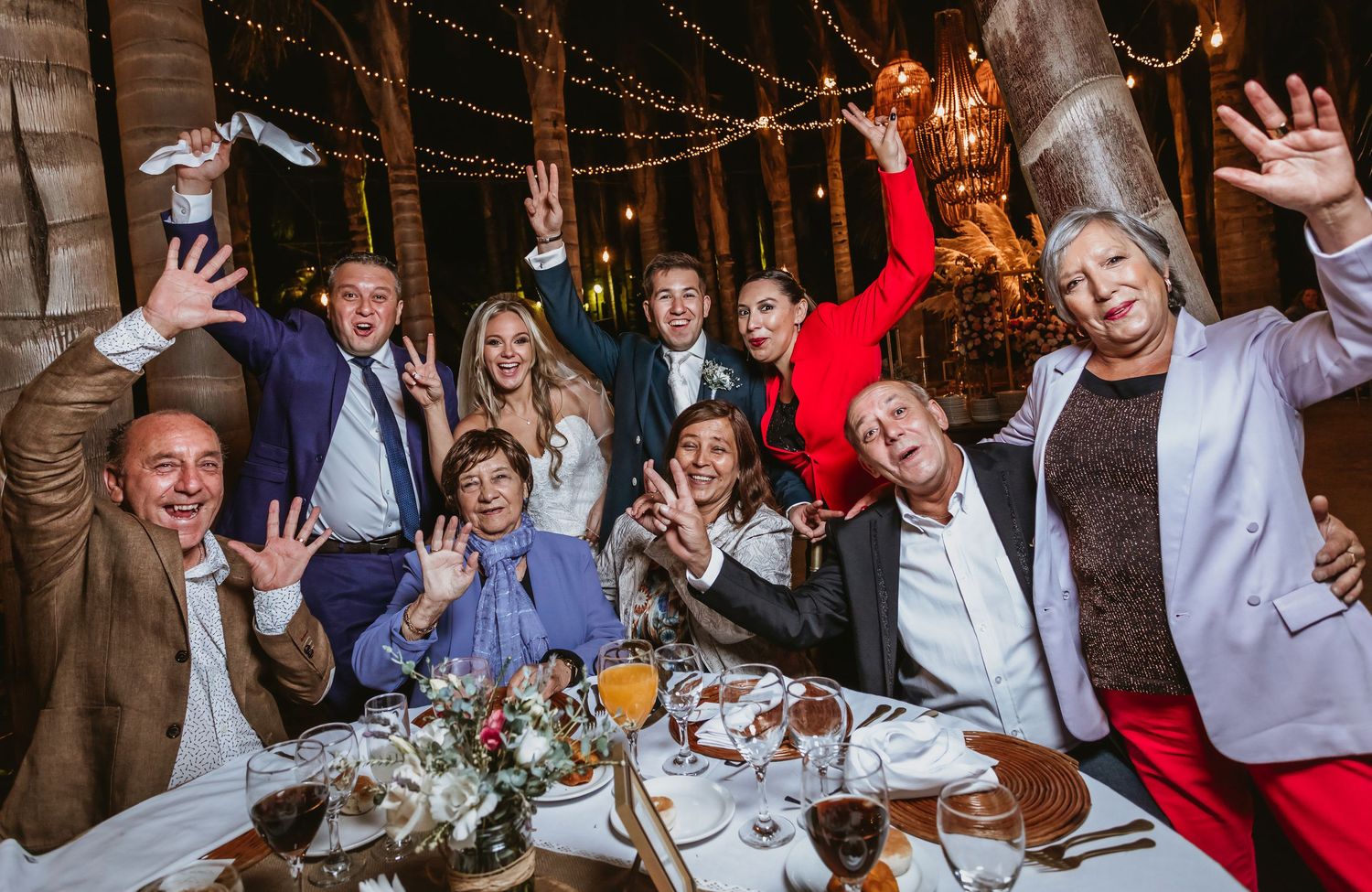 Joyful wedding party group photo at decorated dinner table with string lights at night.