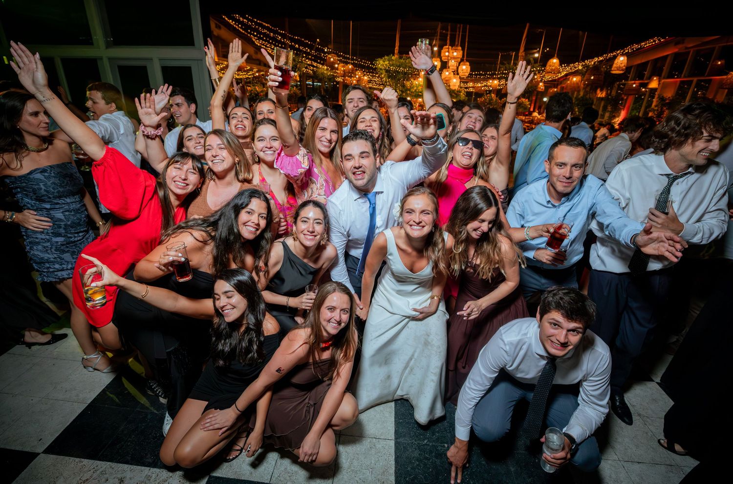 Bride and groom pose with large group of smiling wedding guests for fun outdoor reception photo.