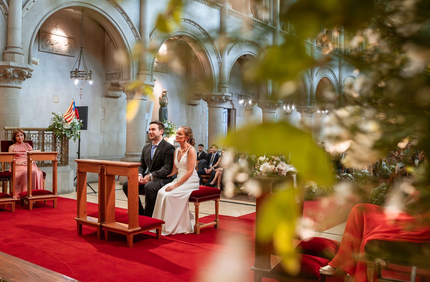 Bride and groom sit together at church pew during wedding ceremony framed by foliage.