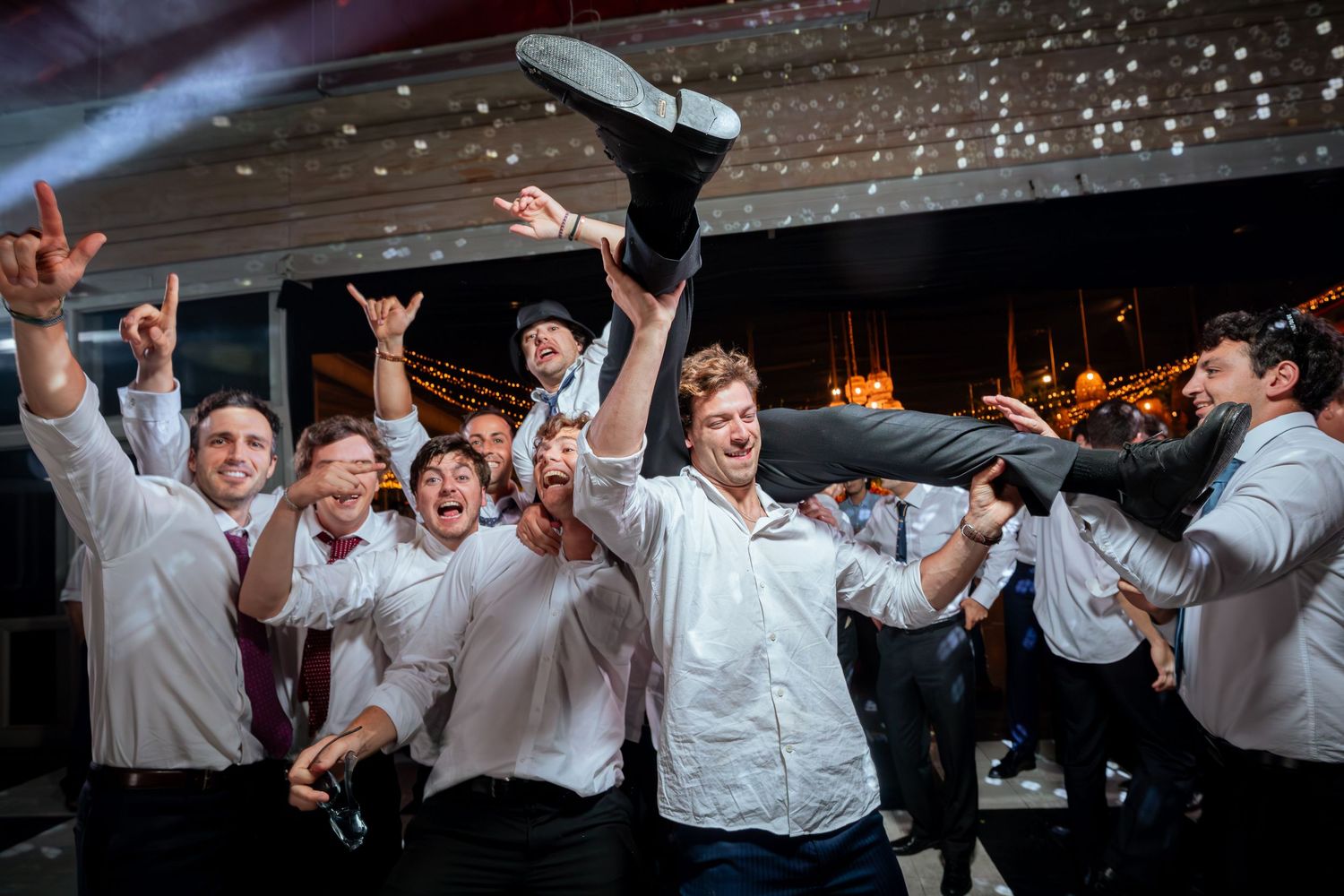 Excited groomsmen in white shirts lift man into air at wedding reception.