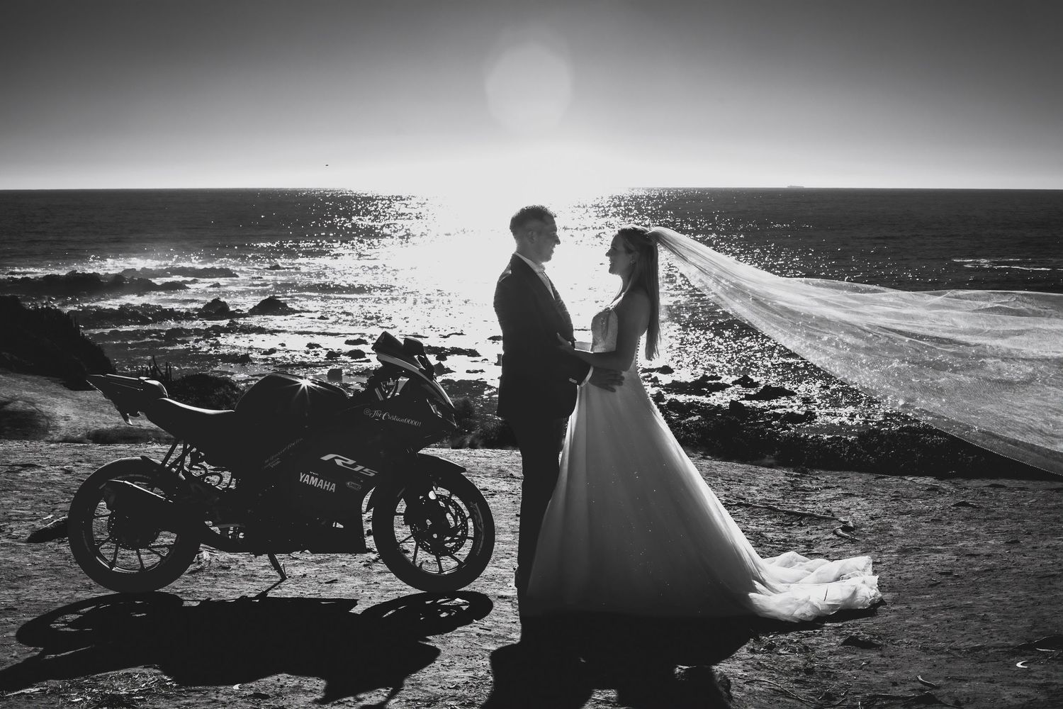 Black and white wedding portrait of couple embracing beside a motorcycle with dramatic ocean and veil flowing behind.