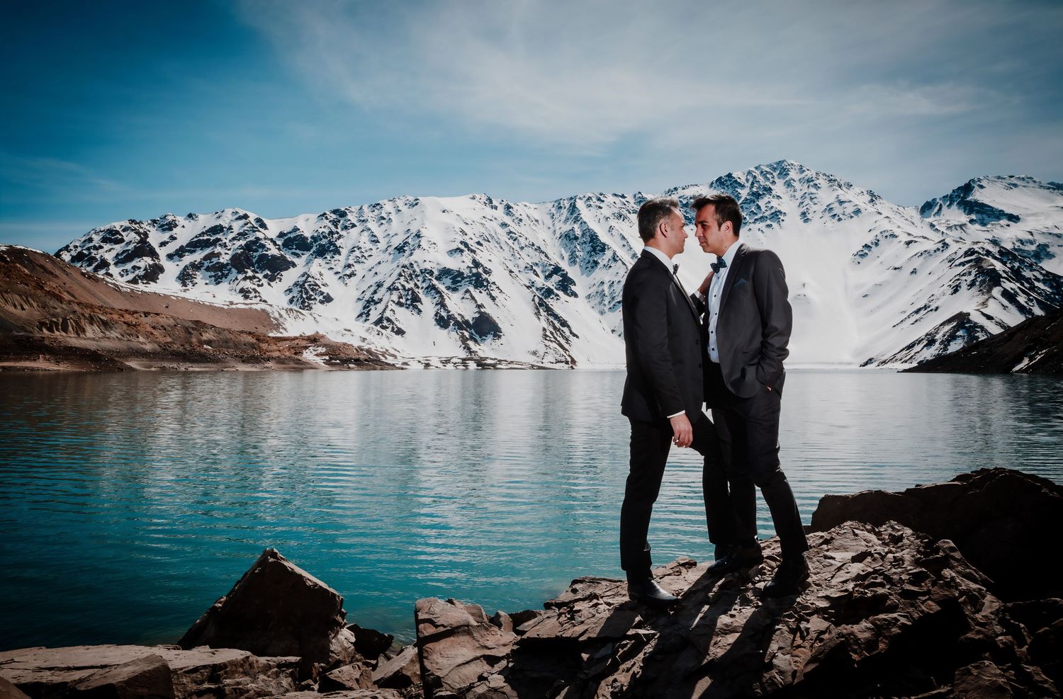 Two grooms share an intimate moment near a stunning turquoise lake surrounded by snowy mountains.