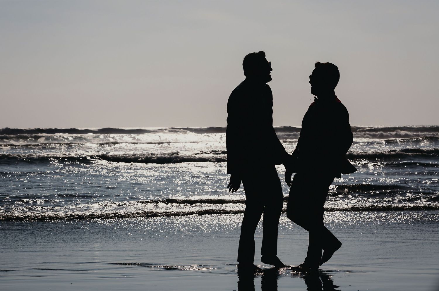 Two grooms silhouetted hand-in-hand walking along a shimmering beach at sunset.