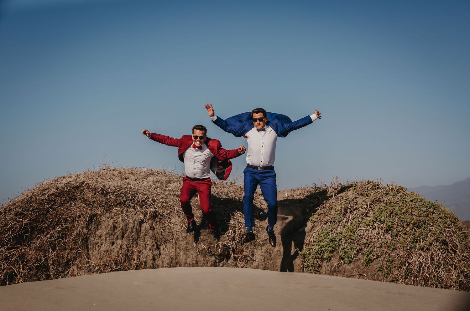 Two grooms in red and blue suits jumping triumphantly on desert dunes under a bright blue sky.