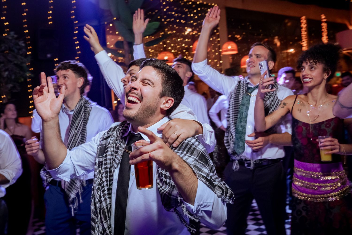 Excited wedding guests dancing and celebrating on the dance floor, man in foreground holding a drink.